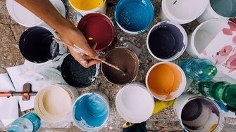 Picture of different buckets with all different types of colors of acrylic paint. You see a hand in the picture, holding a paint brush, reaching out for the brown paint. 