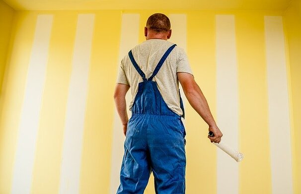 Picture of a man in overall looking at a newly painted yellow wall. This picture is taken from behind this man, so you are looking to his back. 