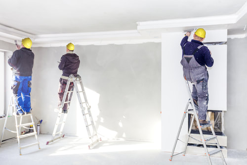 Picture of three men working on a drywall installation. They are all standing on a ladder because they are putting drywall mud towards the ceiling. They are wearing the necessary safety gear like helmets and overalls. 